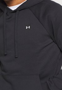 Felpa nera Under Armour con cappuccio, cordini e tasca a marsupio indossata sopra una maglietta bianca, con il logo del marchio visibile sul petto.