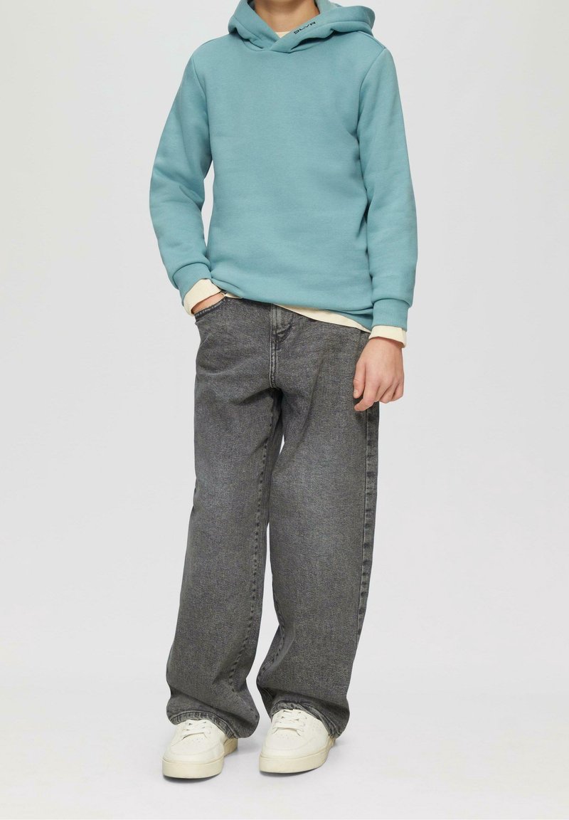 Enfant portant un sweat à capuche bleu clair, un jean large gris foncé, des baskets blanches et une chemise beige en dessous, les mains détendues.