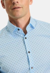 Hellblaues Hemd mit Knöpfen und einem kleinen, wiederholten geometrischen Muster in Dunkelrot. Verfügt über einen spitzen Kragen und dunkle Knöpfe. Glatte Textur.