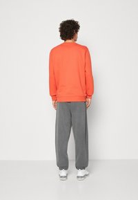 Felpa arancione a maniche lunghe, pantaloni di velluto a coste grigi con polsini elastici e sneaker bianche con dettagli argentati, viste da dietro.
