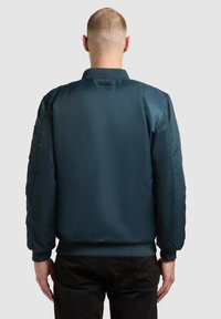 Námořnická modrá bomber bunda s lesklým povrchem, žebrovanými manžetami a lemem a polstrovanými rameny. Disponuje minimalistickým designem a žádnými viditelnými logy.