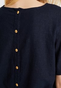 Námořnická modrá lněná blůza s kulatým výstřihem a dřevěnými knoflíky na zádech. Krátké rukávy a jemná textura.