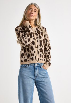Pimpernøtt cardigan i myk beige ull, med knappelukking, ribbestrikkede mansjetter, og en avslappet passform. Kombinert med lyseblå jeans.
