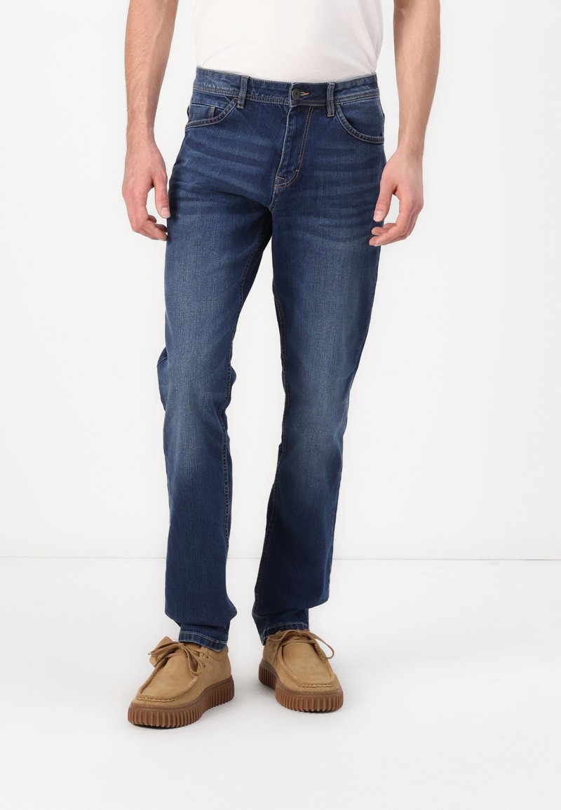 Homme portant un jean droit bleu foncé avec des détails délavés, des chaussures beiges à lacets et une chemise blanche rentrée, debout sur un sol blanc.