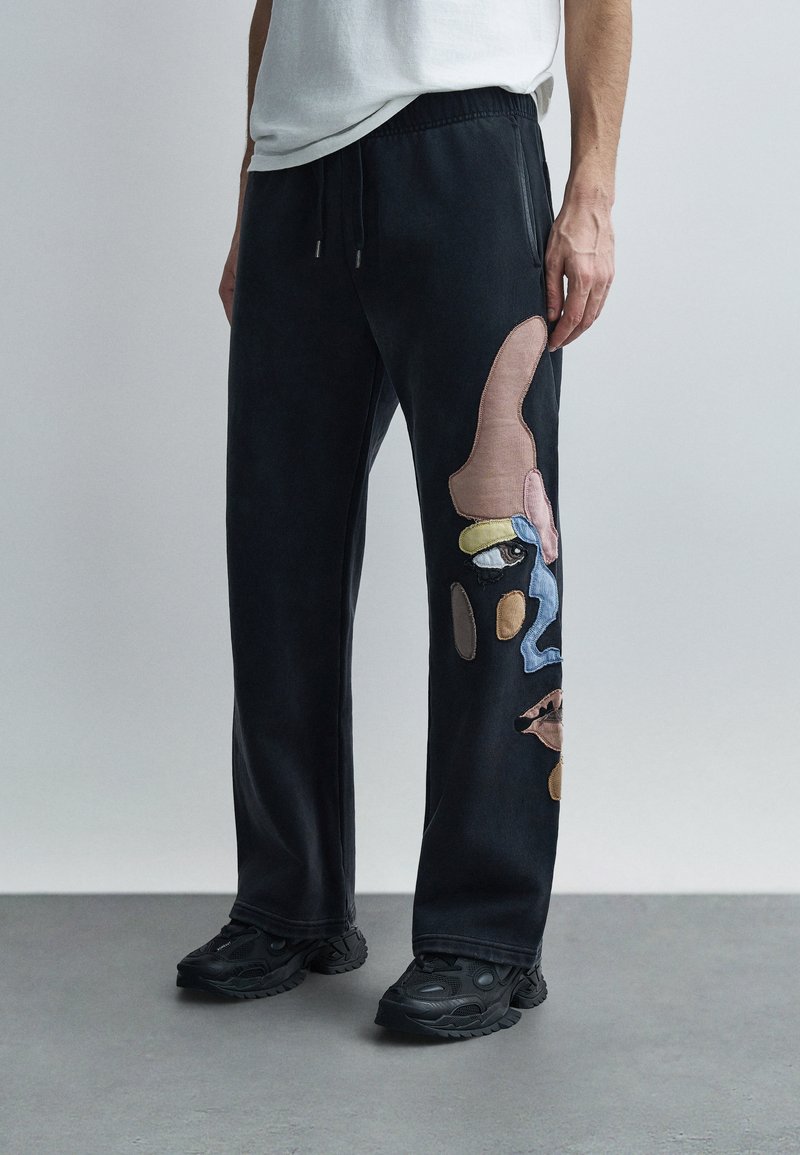 Μαύρο παντελόνι φόρμας με πολύχρωμες αφηρημένες εφαρμογές στο αριστερό πόδι. Κατασκευασμένο από μαλακό ύφασμα με ελαστική μέση και ελαστικό τελείωμα.