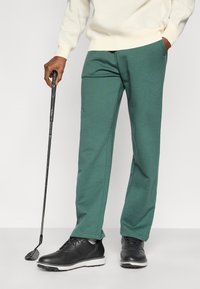 Πράσινο, απαλό ύφασμα φόρμας με άνετη εφαρμογή, σε συνδυασμό με μαύρα, λείωσε δερμάτινα παπούτσια και ένα γκολφ ρόπαλο στο χέρι ενός ατόμου.