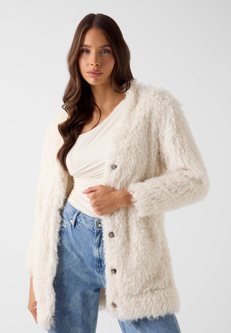 Pluizige witte cardigan met een textuurrijke afwerking, voorzien van grote knopen, lange mouwen en een open voorkant, gecombineerd met een licht topje en blauwe spijkerbroek.