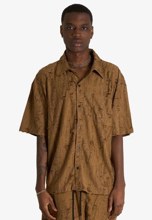 Giovane con capelli corti che indossa una camicia marrone testurizzata a maniche corte con bottoni e pantaloni abbinati su uno sfondo semplice.