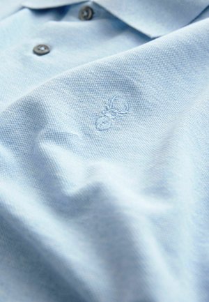 Svetlomodrá polokošeľa vyrobená z textúrovanej látky, ktorá má dva tmavé gombíky a vyšívané logo pavúka na ľavej strane hrude.