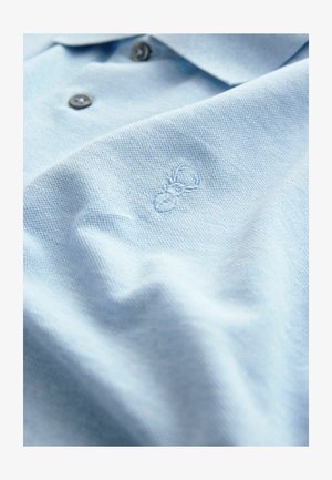 Svetlomodrá polokošeľa vyrobená z textúrovanej látky, ktorá má dva tmavé gombíky a vyšívané logo pavúka na ľavej strane hrude.