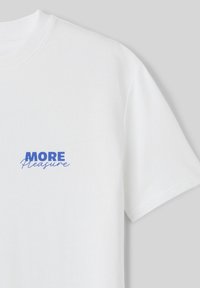 T-shirt en coton blanc avec un design graphique en bleu sur la poitrine gauche, portant l'inscription "PLUS de Plaisir". Manches courtes.