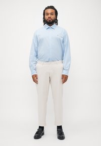 Camisa de botón azul claro con pequeños puntos blancos, pantalones beig y mocasines negros. El modelo está de pie con las manos entrelazadas al frente.