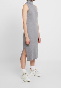 Grå ribbad stickad klänning med hög krage, sidofickor och ärmlös design, ihop med vita sneakers och ankelsockor.