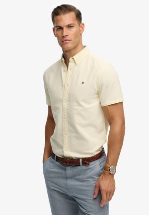 Hombre con camisa crema de manga corta abotonada, pantalones azul claro, cinturón trenzado marrón y reloj de pulsera, de pie con las manos en los bolsillos.