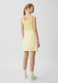 Junge Frau mit hellbraunem Haar in einem Pferdeschwanz, die ein gelbes Tanktop, einen hellgelben Rock, gelbe Socken und weiß-gelbe Turnschuhe trägt und von hinten schaut.