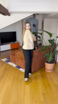 Svetlo bež kratek rokav zgornji del obleke, črne hlače in srebrni supergi, stoječi v sodobni sobi z rjavim kordurovim kavčem in lončnico.