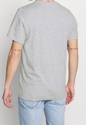 Person trägt ein schlichtes hellgraues T-Shirt mit kurzen Ärmeln und hellblaue Jeans, von hinten betrachtet, mit Tätowierungen von Stacheldraht auf beiden Oberarmen.