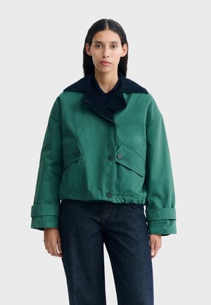 Femme aux cheveux raides et foncés portant une veste verte avec de grands revers et des sangles aux poignets, associée à un jean bleu foncé, sur un fond uni.