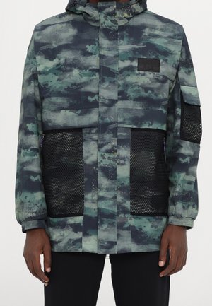 Veste à motif camouflage avec poches en filet noir à l’avant et sur la manche, capuche et poignets élastiques portée par une personne debout avec les bras détendus.