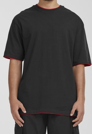 Mężczyzna w luźnym czarnym T-shircie z czerwonym obszyciem na kołnierzu, rękawach i dolnym brzegu, stojący na tle jednolitego jasnego tła.