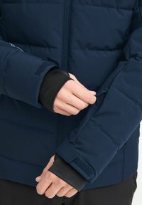 Jūros karines spalvos paminkštintas džemperis su juodais elastingais rankogaliais; turi užtrauktuko kišenę ir Velcro užsegimą ant rankovės, leidžiantį pritaikyti dydį.