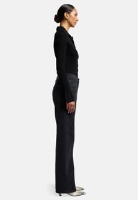 Top a maniche lunghe nero a coste abbinato a jeans scuri a vita alta e scarpe con tacco a punta in stampa pitone. La modella è in profilo contro uno sfondo bianco.