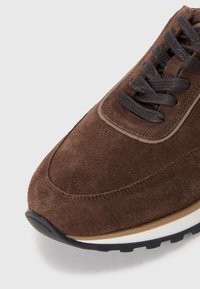 Brown suede sneaker with dark laces, a rounded toe, contrasting light accents, and a textured sole for grip and stability.