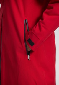 Veste imperméable rouge avec une poche latérale zippée, des détails de fermeture éclair noirs et un poignet à fermeture velcro. Le tissu a une texture lisse.