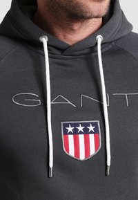 Felpa grigia con cordini bianchi, caratterizzata da un grande scudo ricamato con elementi rossi, bianchi e blu e la parola "GANT" in grigio.