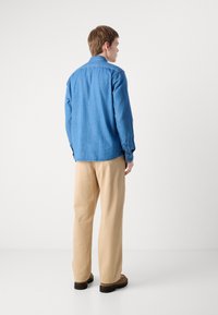 Camicia di denim con maniche lunghe, colletto a punta e taglio dritto, di colore blu medio. Abbinata a pantaloni beiges a gamba larga e scarpe marroni.
