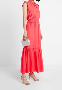 Korallenfarbenes Maxikleid mit Rüschenärmeln, taillierter Taille und gestuftem Rock. Begleitet von einer kleinen runden silbernen Handtasche und nudefarbenen Sandalen.