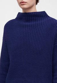 Personne portant un pull épais en tricot bleu foncé avec un col montant large et côtelé, montrée des épaules jusqu'à juste en dessous du nez.