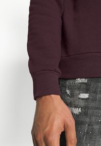 Mano destra di una persona che indossa un maglione a maniche lunghe color marsala e jeans neri strappati.