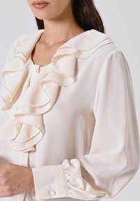 Donna che indossa una blusa cremosa con un ampio colletto a ruches e maniche a sbuffo, abbottonata ai polsi, su uno sfondo neutro.