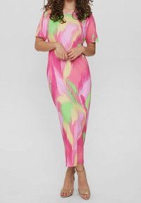 Femme portant une robe midi à motifs abstraits colorés rose, vert et jaune, avec des manches courtes et des talons nus à bride de cheville, les mains jointes.