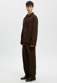 Brązowa bawełniana kurtka z zapięciem na guziki i kieszenią na piersi, zestawiona z pasującymi szerokimi spodniami. Czarne buty dopełniają stylizację.
