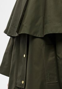 Casaco impermeável verde-oliva com design em camadas, fechos de pressão dourados, bolso frontal e marcação subtil na lateral.