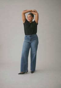 Melnas īss piedurkņu T-krekls kopā ar augstu jostasvietu un plašām samta zilām džinsu biksēm. Modelis valkā melnas smailas papēžu kurpes, stāvot pret neitrālu fonu.