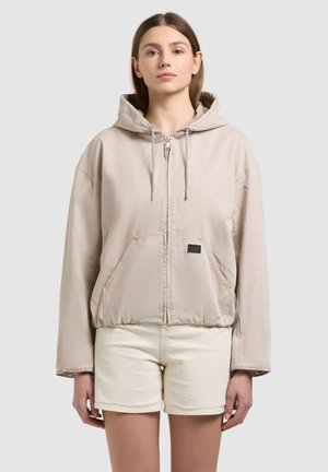 Fiatal nő bézs színű cipzáras kapucnis dzsekit és törtfehér rövidnadrágot visel, egy egyszínű világos háttér előtt áll.