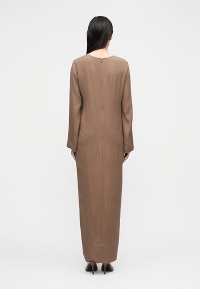 Γυναίκα με μακριά σκούρα μαλλιά, ντυμένη με καφέ φόρεμα μακριάς γραμμής και μακριά μανίκια, και καφέ ψηλοτάκουνα παπούτσια, στέκεται πλάτη προς τα εμάς.