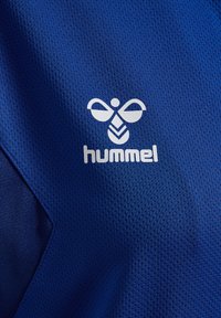 Maglietta sportiva in rete blu con superficie testurizzata, con un significativo logo Hummel bianco posizionato al centro.