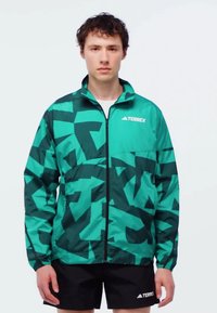 Grön och mörk teal jacka med abstrakt mönster, hel dragkedja, ståkrage och långa ärmar, kombinerad med svarta shorts med logotyp.