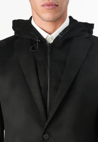 Schwarze Hybridjacke mit Kapuze, Reißverschluss vorne und taillierten Revers. Hergestellt aus glattem Stoff mit einem subtilen Glanz. Ein-Knopf-Verschluss.