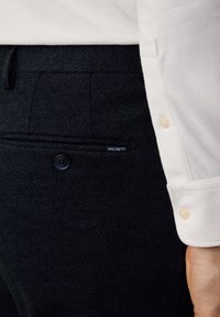Marineblauwe herringbone-gepatroneerde broek met een achterzak en knoopsluiting, voorzien van een blauwe label met de tekst "HACKETT." Witte mouw van een shirt zichtbaar.