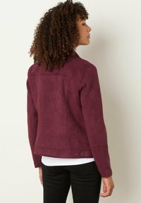 Veste en daim bordeaux avec une silhouette ajustée, présentant une texture lisse, des détails cousus et une fermeture à pression sur l'ourlet.