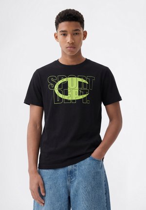 Jeune homme aux cheveux courts et bouclés portant un t-shirt noir avec un imprimé jaune "SPORT DEPT." et un jean bleu, debout sur un fond blanc.