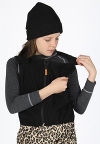 Schwarze, mit Reißverschluss versehene Weste mit Schulterriemen und einem strukturierten Stoff, die über einem grauen Langarmshirt und einer Leopardenmuster-Hose getragen wird.