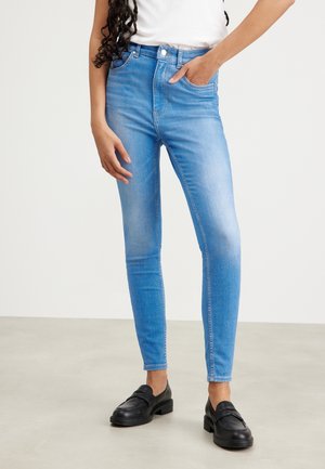 Femme portant un jean skinny bleu clair et des mocassins noirs, la main dans la poche, debout sur un sol gris clair contre un mur blanc uni.