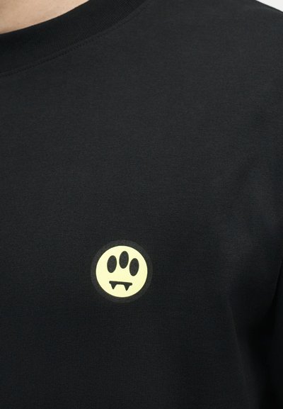 Μαύρο πουκάμισο με μικρό κίτρινο κυκλικό λογότυπο που δείχνει τρία μαύρα ωοειδή πάνω από δύο βρικολακίστικους κυνόδοντες στην περιοχή του στήθους.