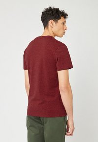 Man die zijwaarts staat, gekleed in een gemêleerd bordeauxkleurig T-shirt met korte mouwen en olijfgroene broek tegen een effen witte achtergrond.